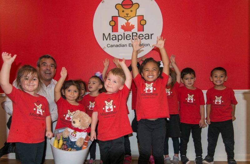 A group of children smiling infront of a Maple Bear Lewisville preschool and daycare sign